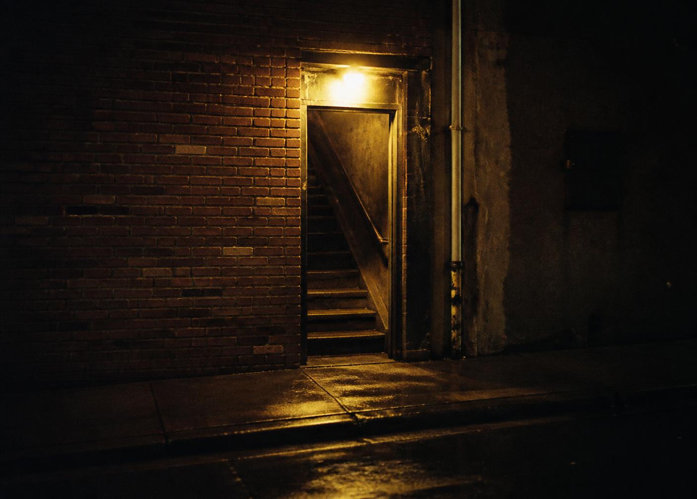 An unmarked doorway and dark stair lit by one warm bulb